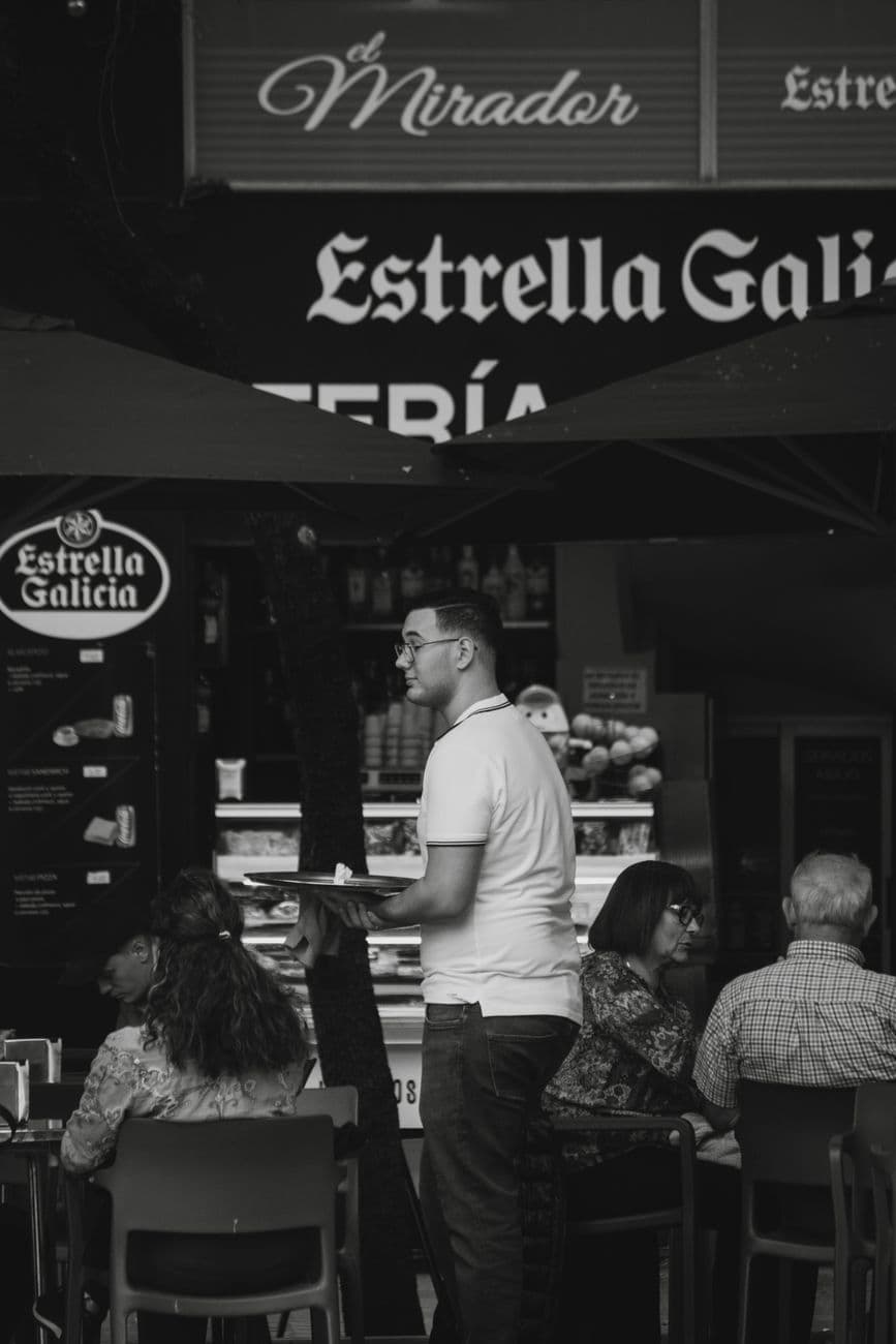 A black and white capture of a waiter serving guests in an outdoor restaurant setting.