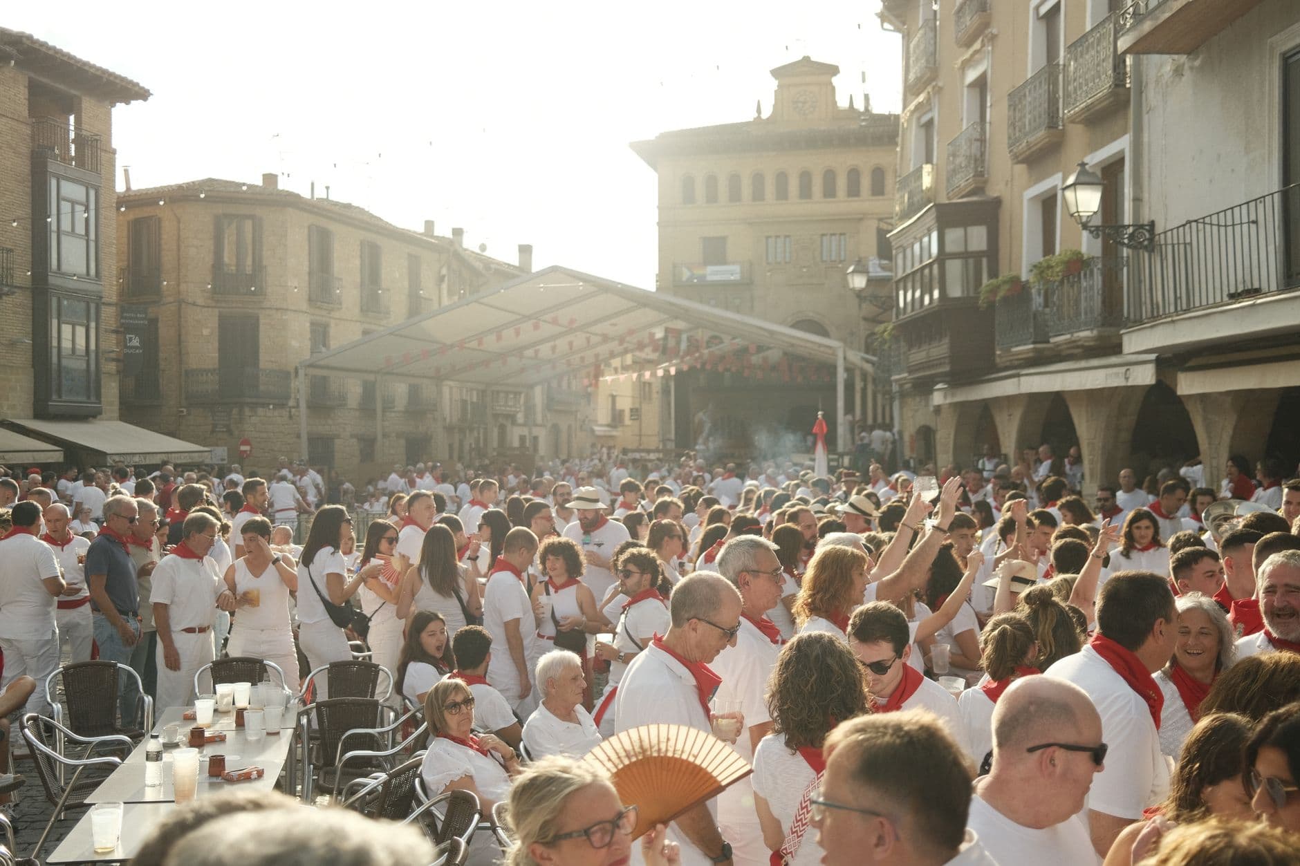Crowded festival in a historic European town square with people in white outfits enjoying sunny weather.