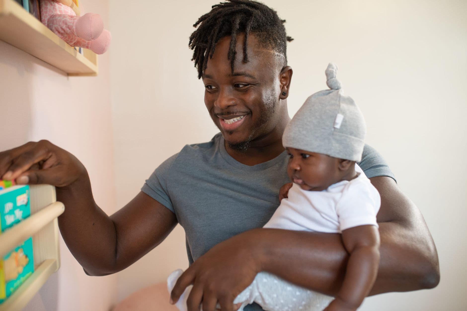 A joyful father with dreadlocks holding his baby indoors, selecting a book.