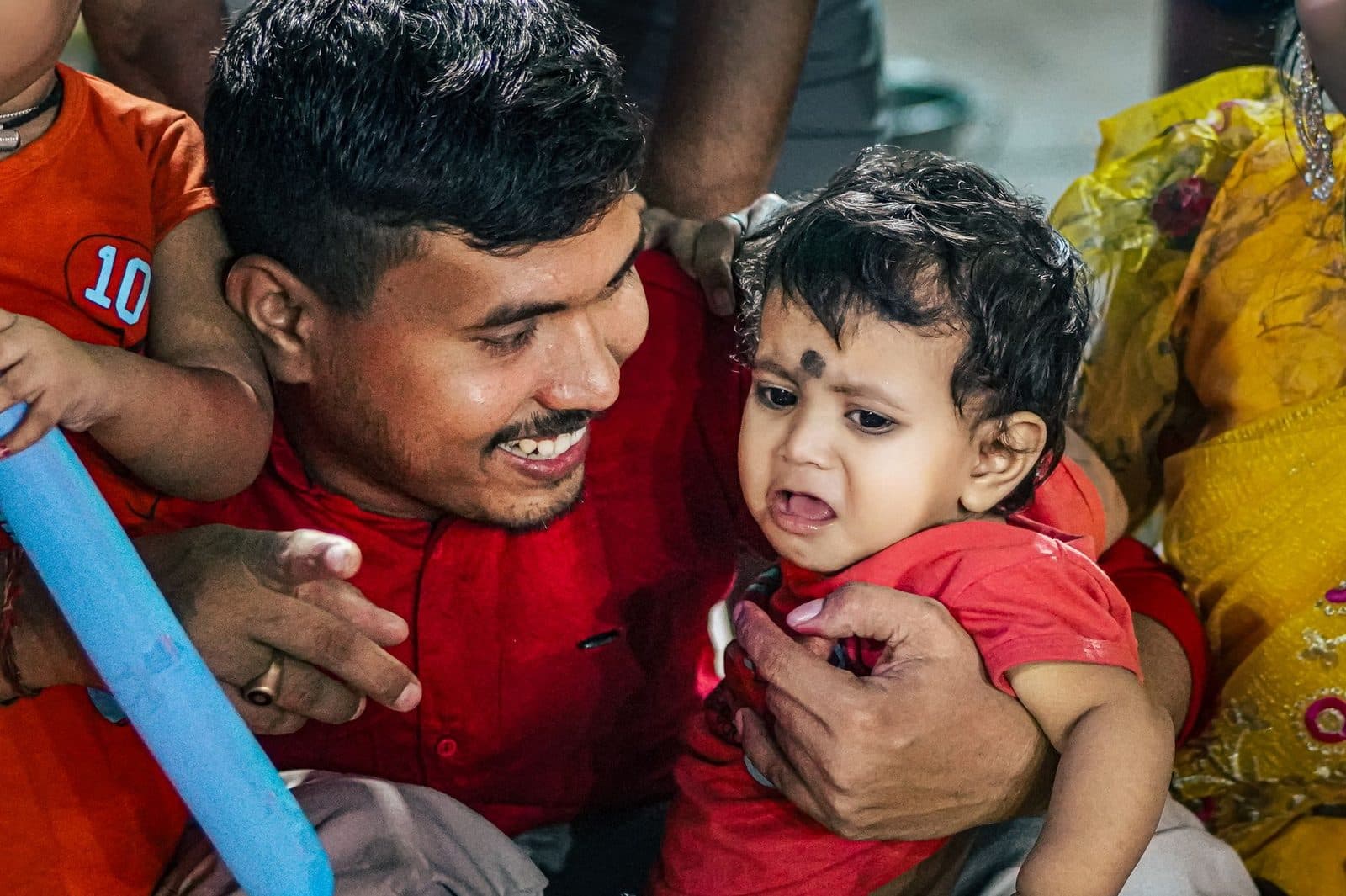 A father lovingly comforts a crying baby at a lively family gathering indoors.