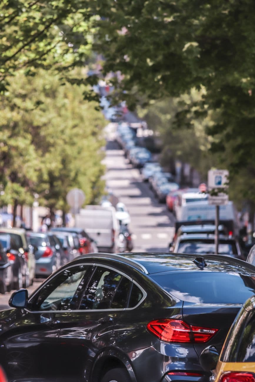 A bustling city street filled with parked cars under a canopy of green trees on a sunny day.