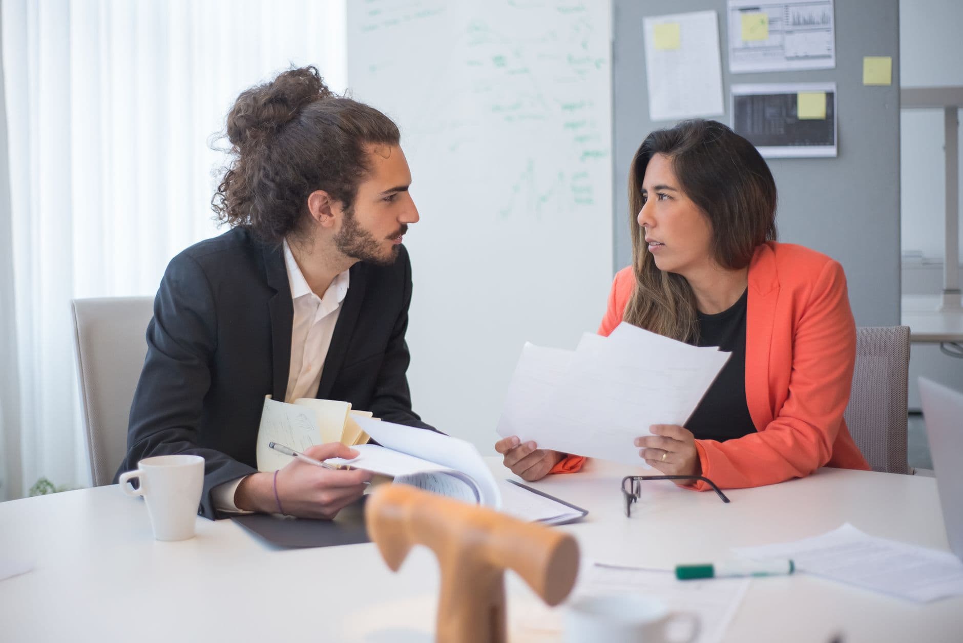 Two colleagues engaged in a discussion at an office table with documents and coffee.