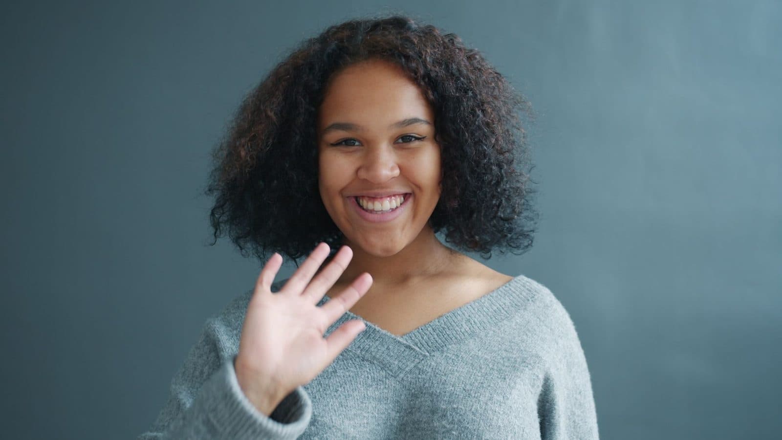 Cheerful woman in grey sweater waving with a smile, indoor setting.