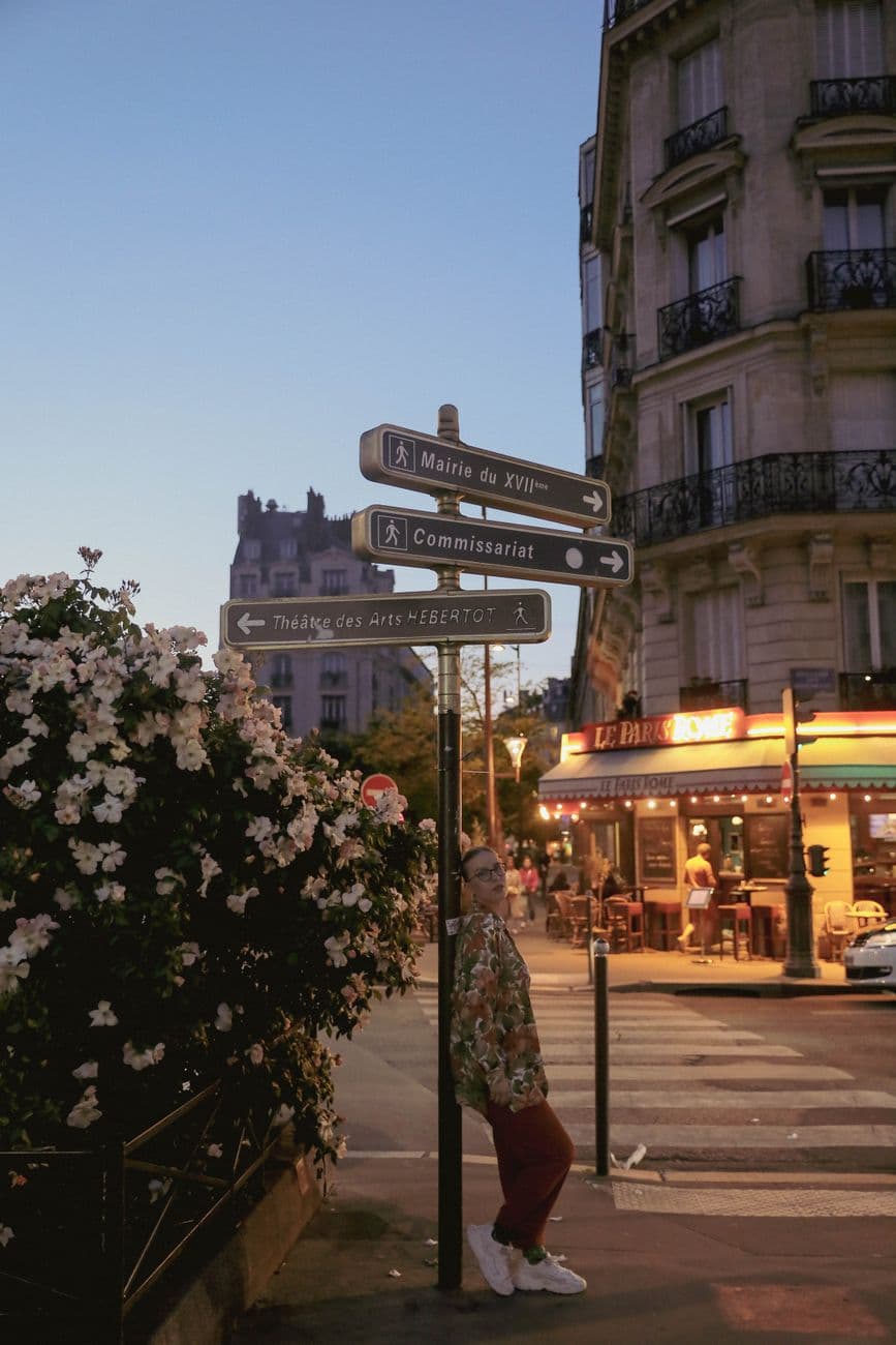 A woman leans on a street sign in Paris, framed by blooming flowers and bustling cafes.