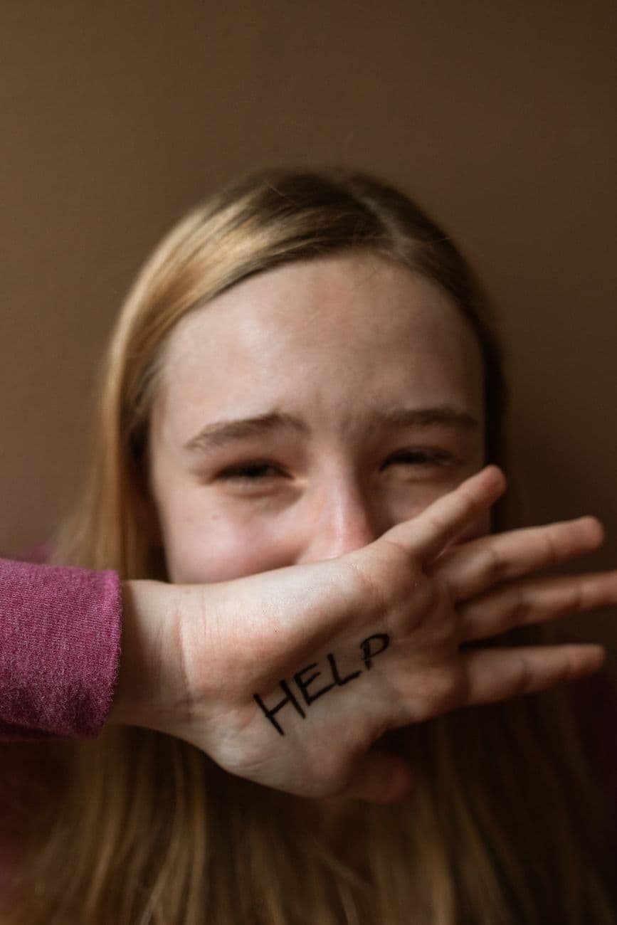 A distressed teenager covering face with 'HELP' written on hand, signifying need for support.