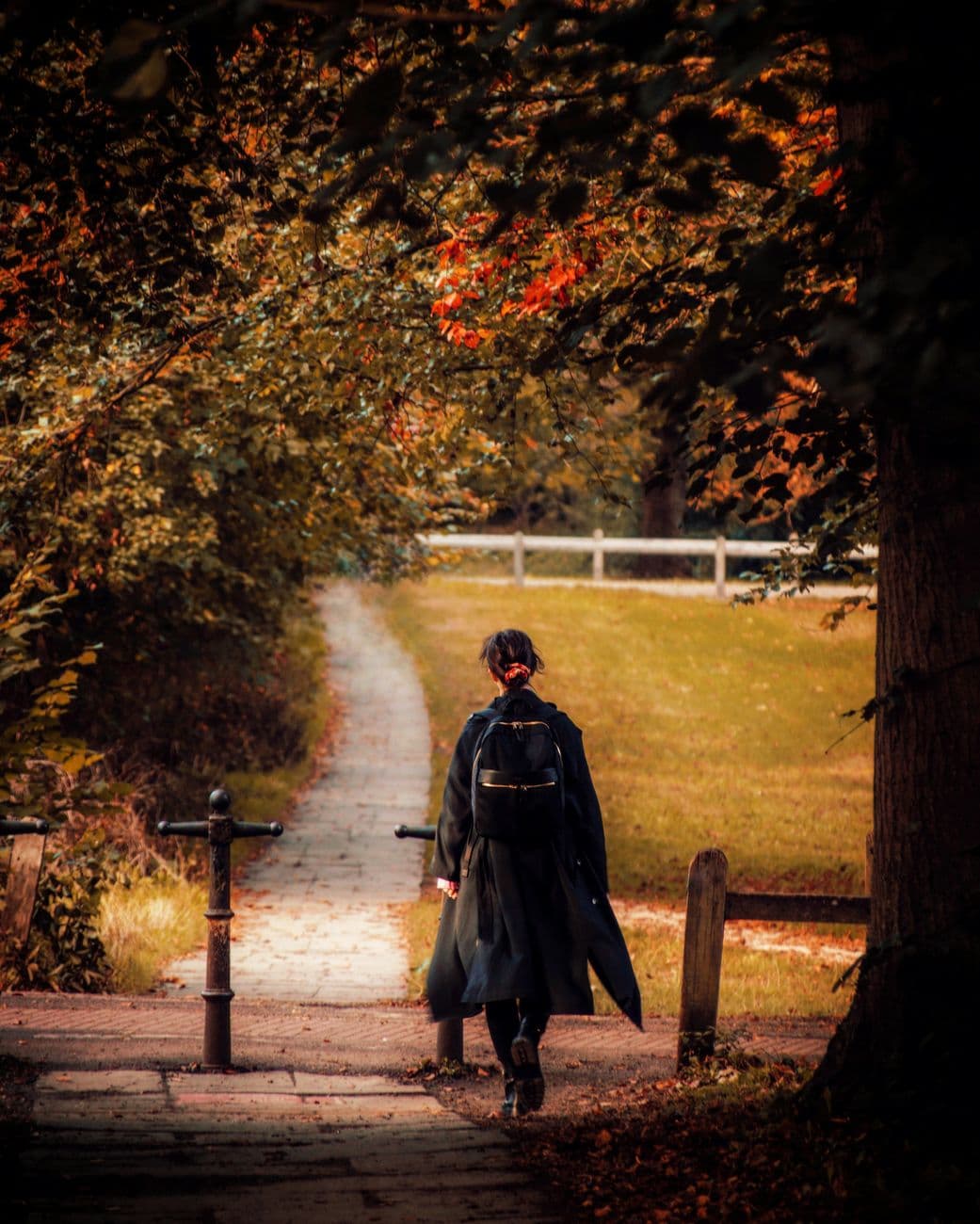 A woman walking along a tree-lined path in a Cambridge park during autumn, showcasing vibrant fall colors.