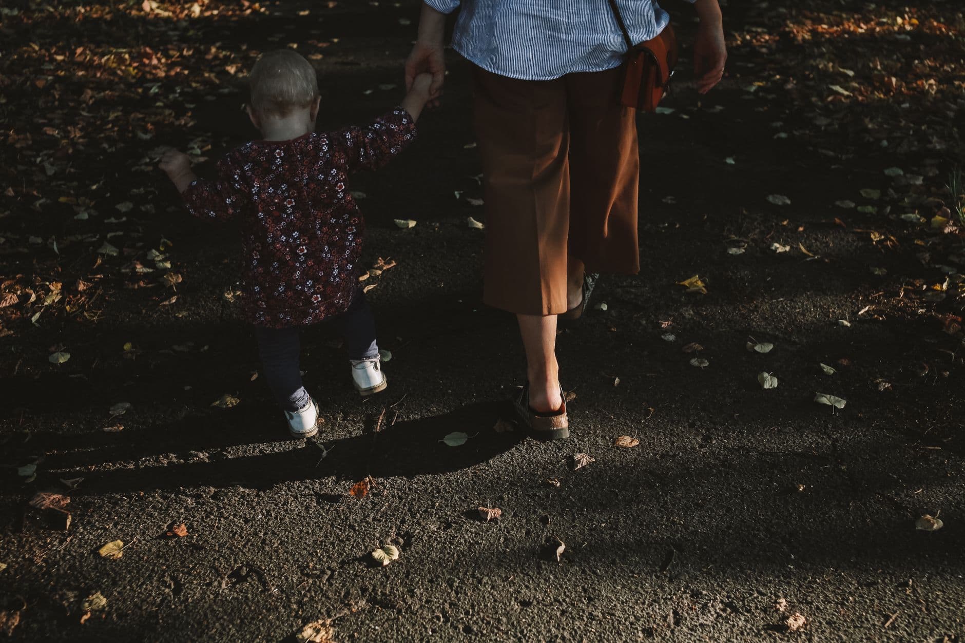A mother and child walking hand in hand on a leaf-strewn path during fall.