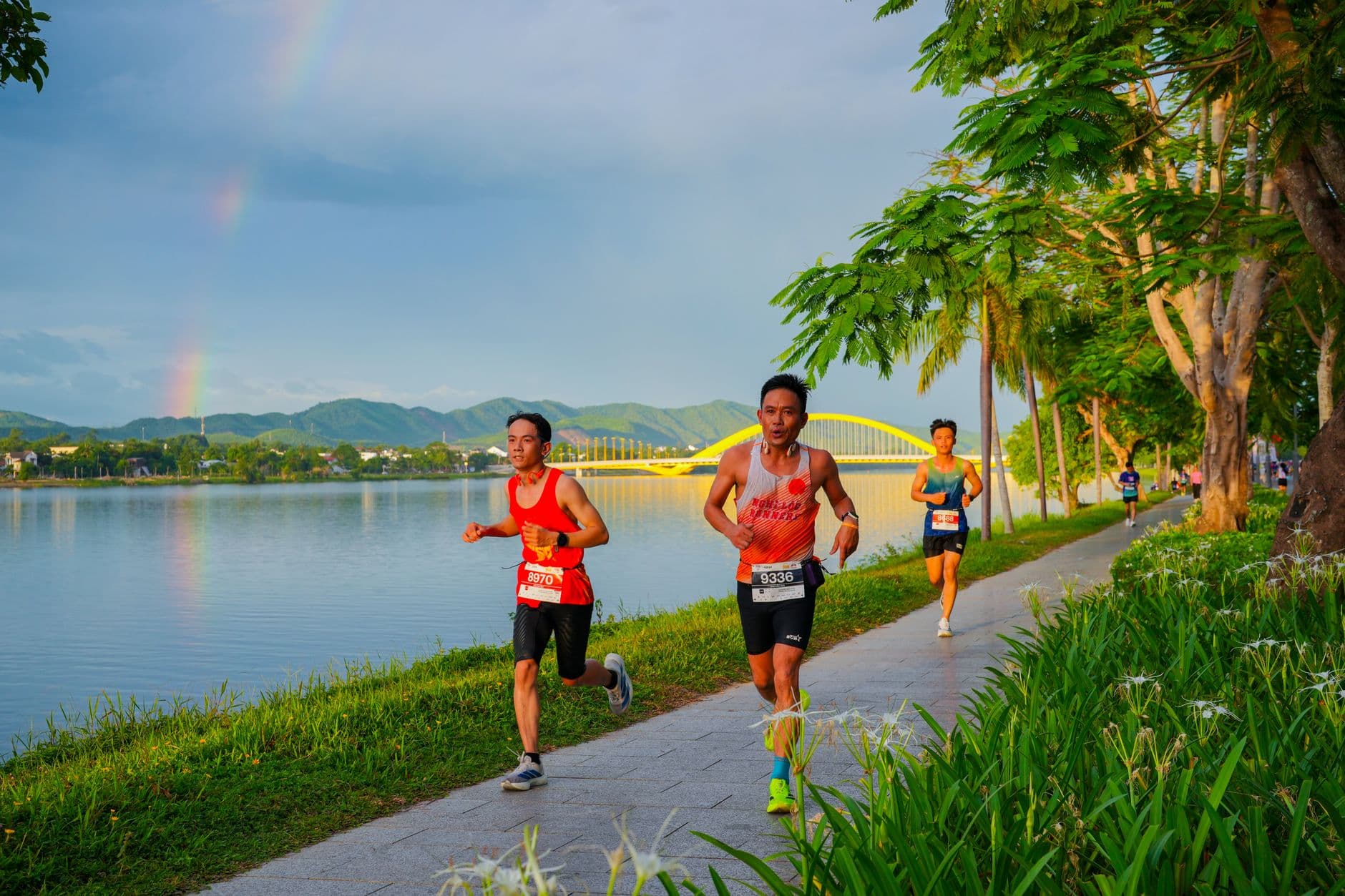 Marathon runners jogging beside a scenic river with a rainbow and iconic bridge.