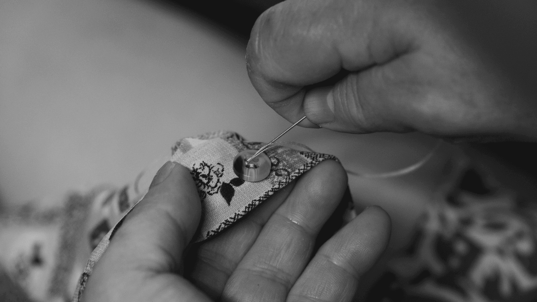 Black and white image of hands sewing a button onto patterned fabric.
