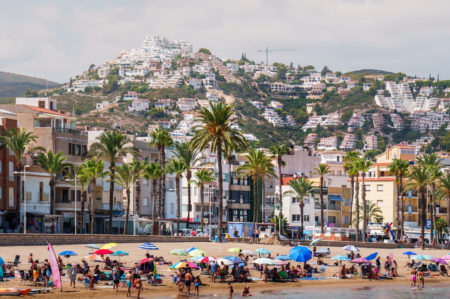 A bustling Spanish beach town with colorful umbrellas, hills, and palm trees.