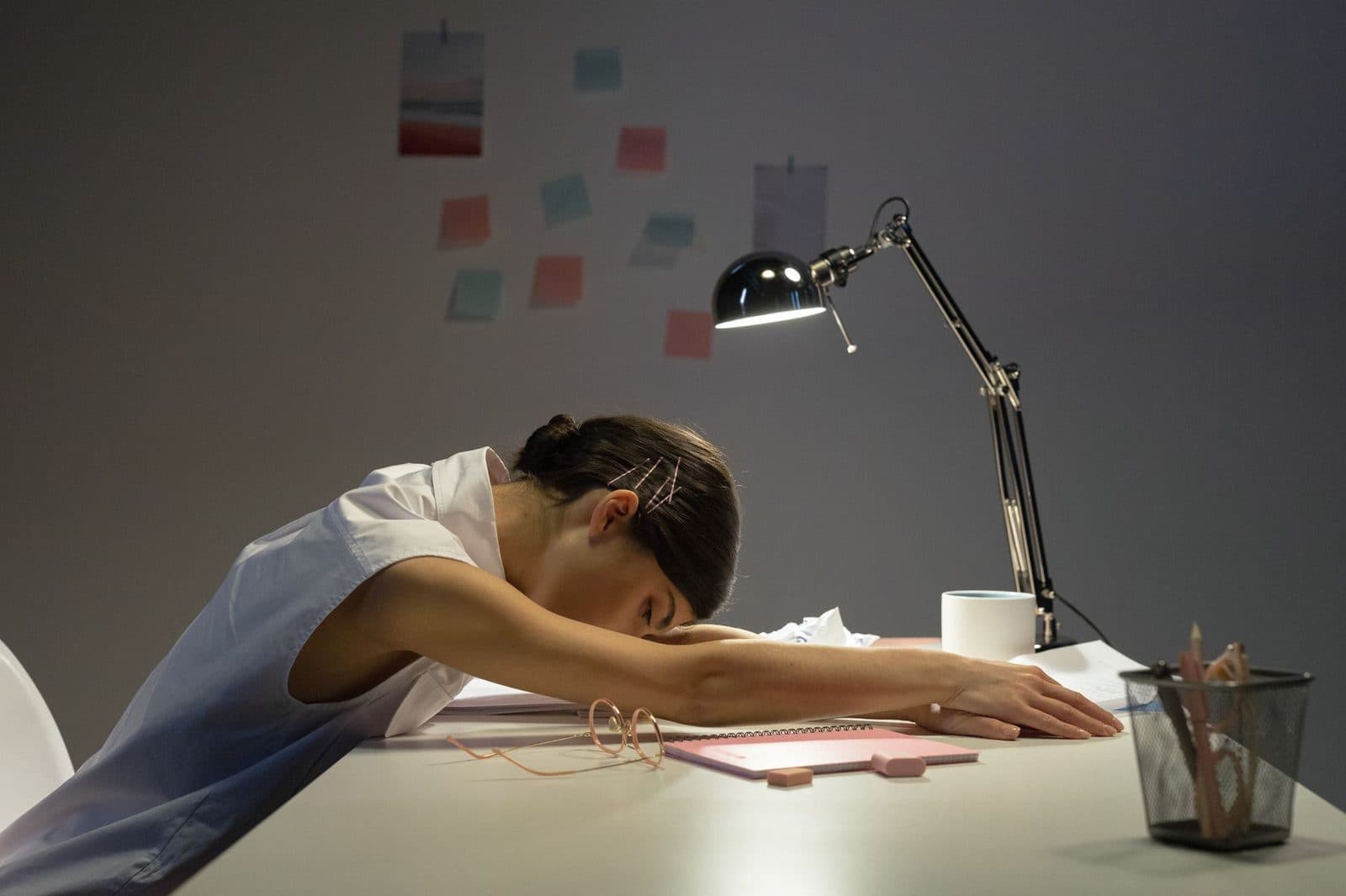 A tired woman rests her head on an office desk under a lamp, symbolizing workplace fatigue.
