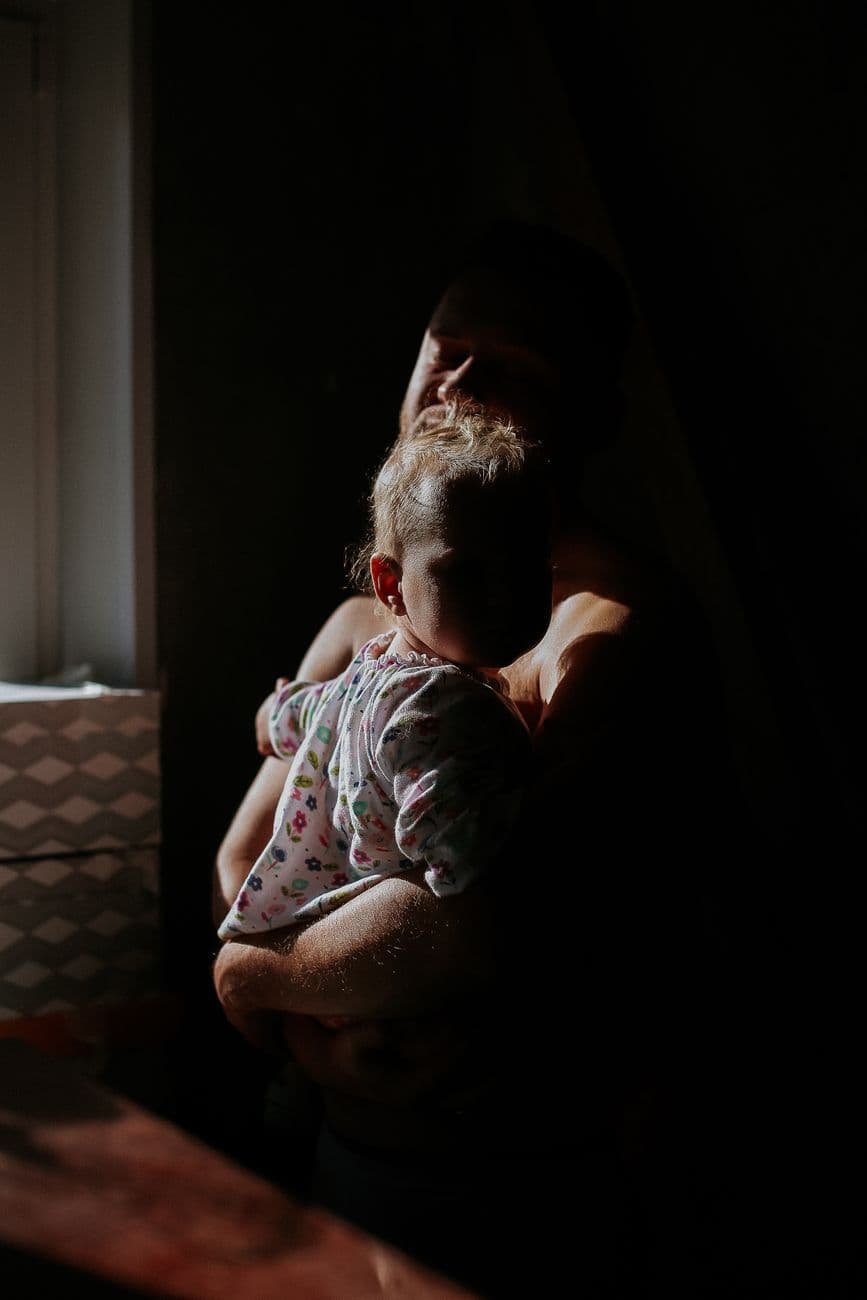 A father lovingly holds his baby by a window in warm morning light, capturing a quiet, tender moment.