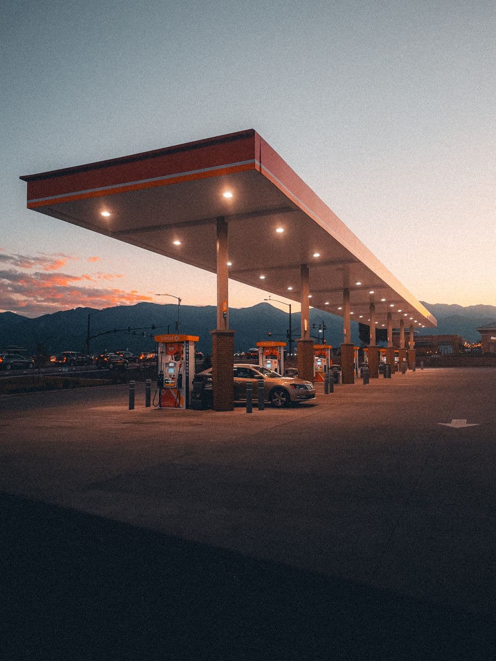 A serene gas station scene at dusk, featuring multiple cars, bright lights, and mountains in the background.