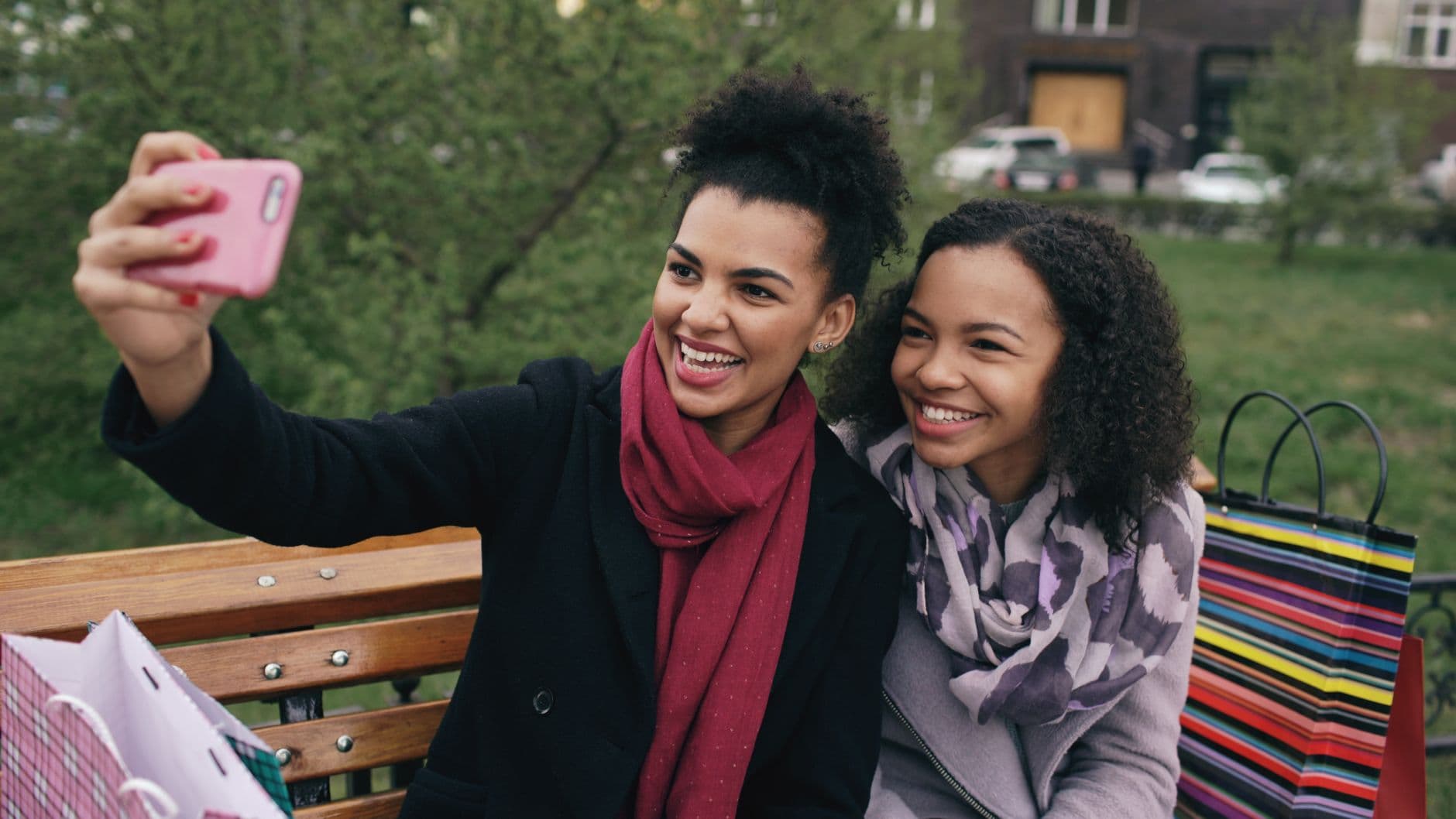 Two smiling friends taking a selfie together on a park bench with shopping bags.