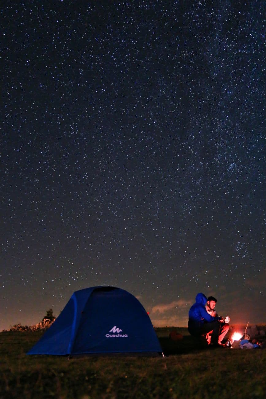 A serene night camping scene with a tent and people by a campfire under a starry sky.
