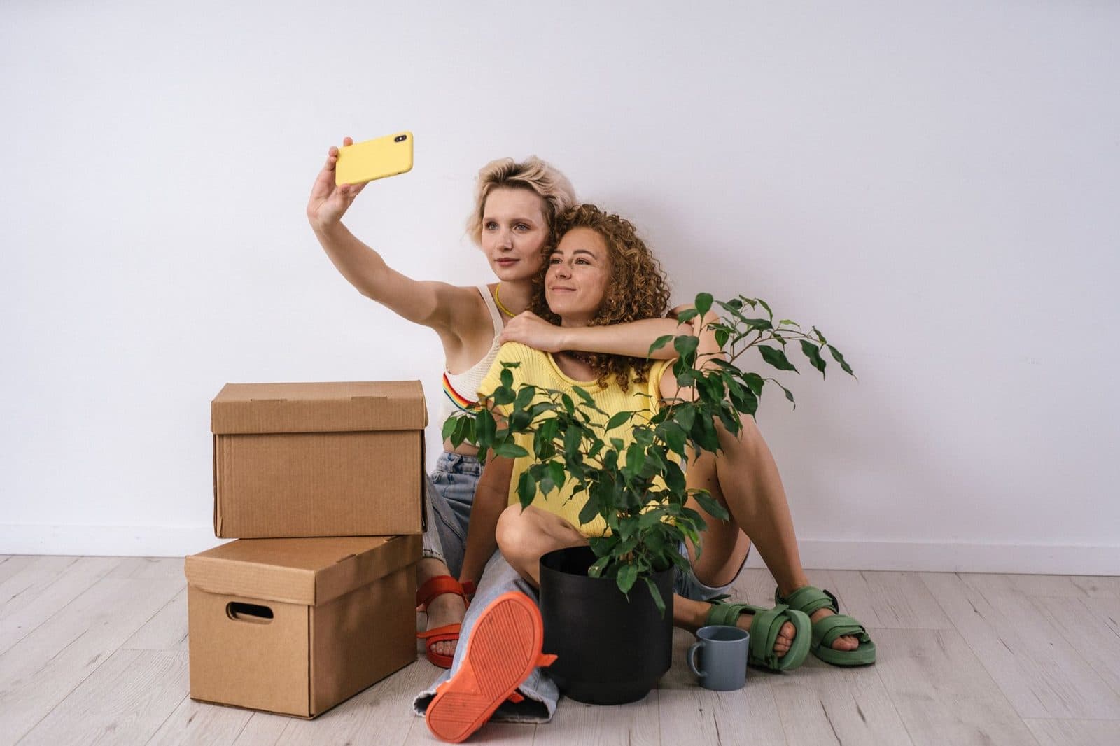 Lesbian couple sitting indoors, embracing and capturing a selfie with a smartphone among boxes and plants.
