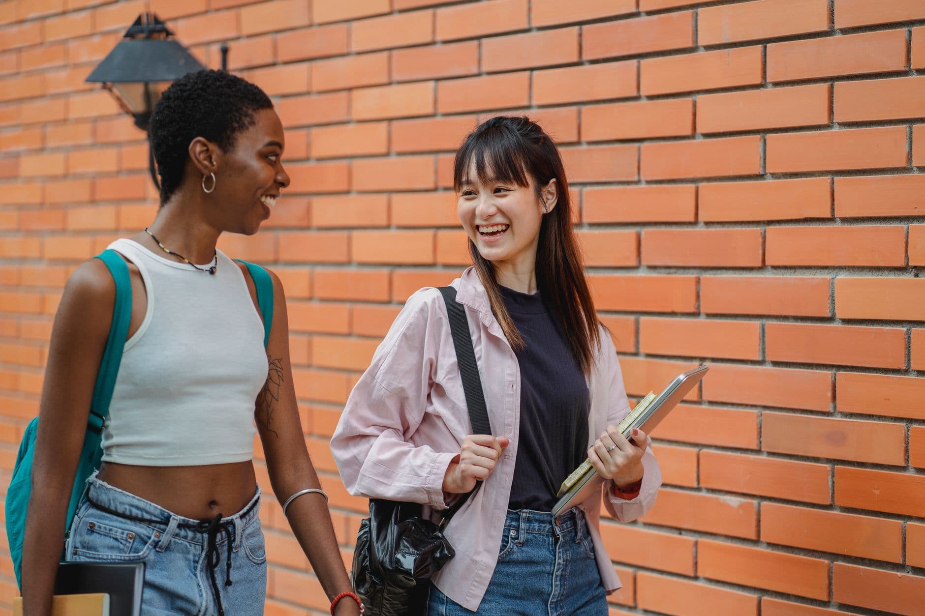 Cheerful multiracial female students wearing casual clothes carrying backpack and notebooks near college brick wall while discussing plans and looking at each other with smiles