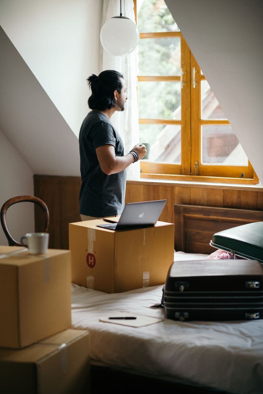 Side view of young ethnic male owner of newly bought house with cup of coffee in bedroom full of boxes and suitcases standing and looking at neighboring houses through window