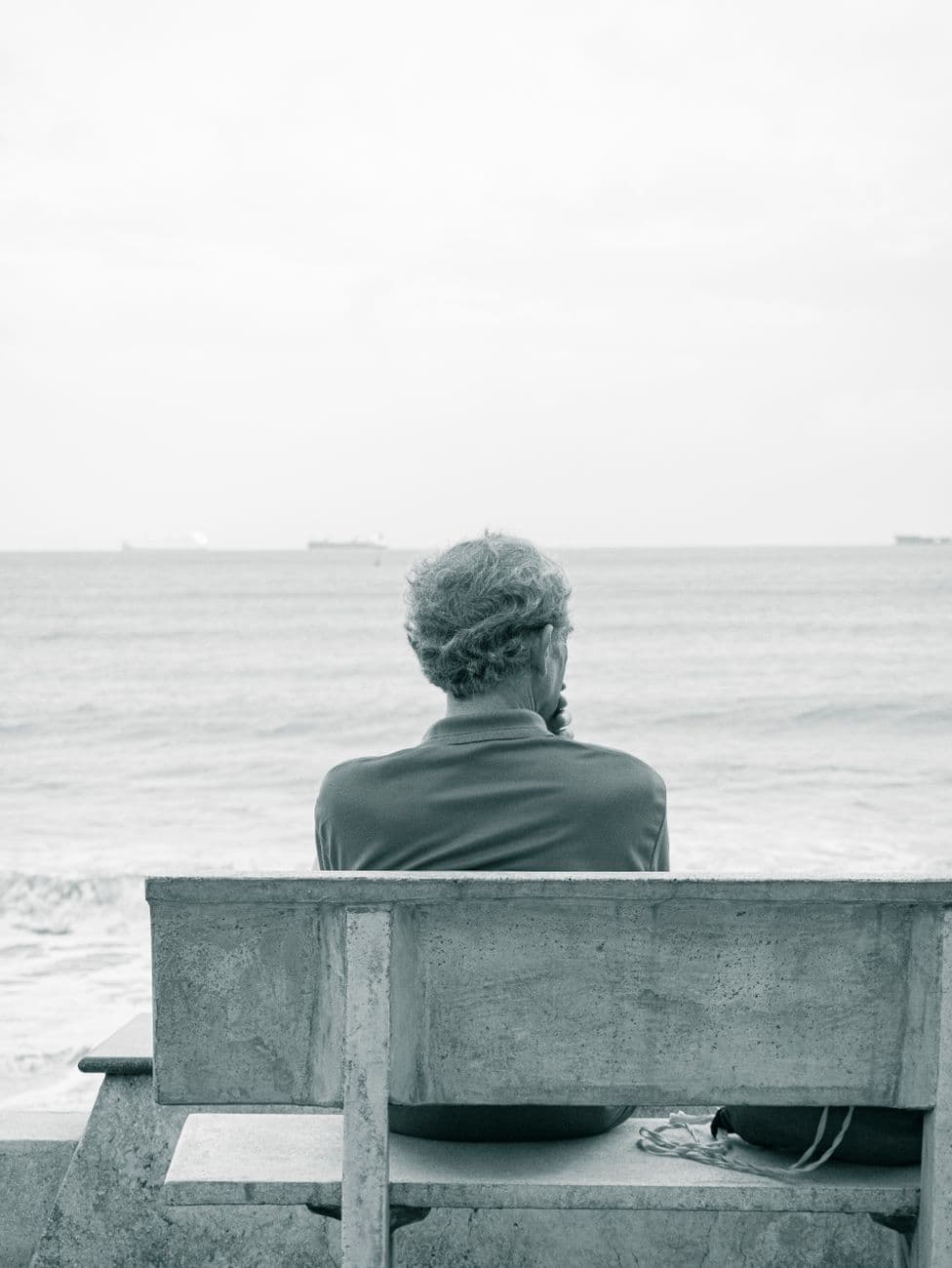 A man sits alone on a bench facing the calm sea, lost in thought.