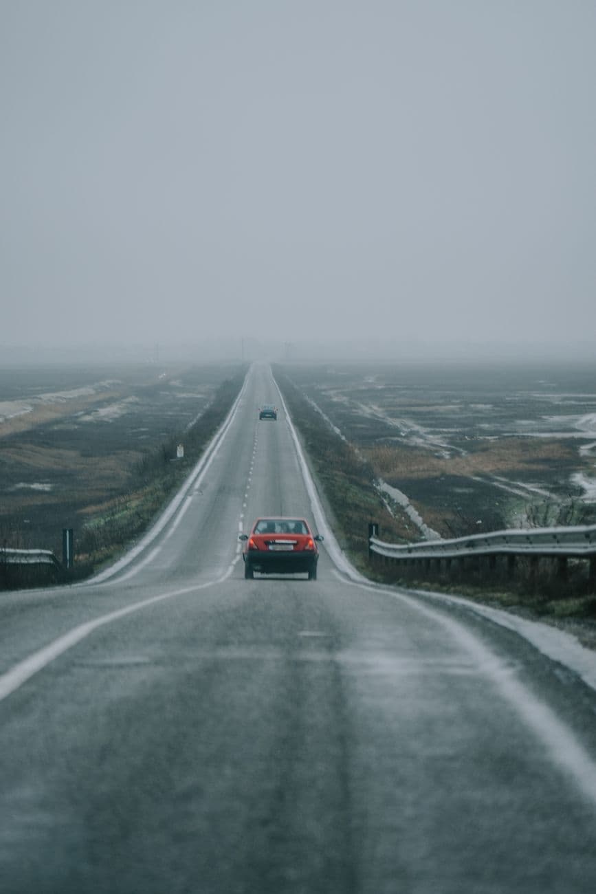 A red car driving on a desolate highway under gloomy skies.