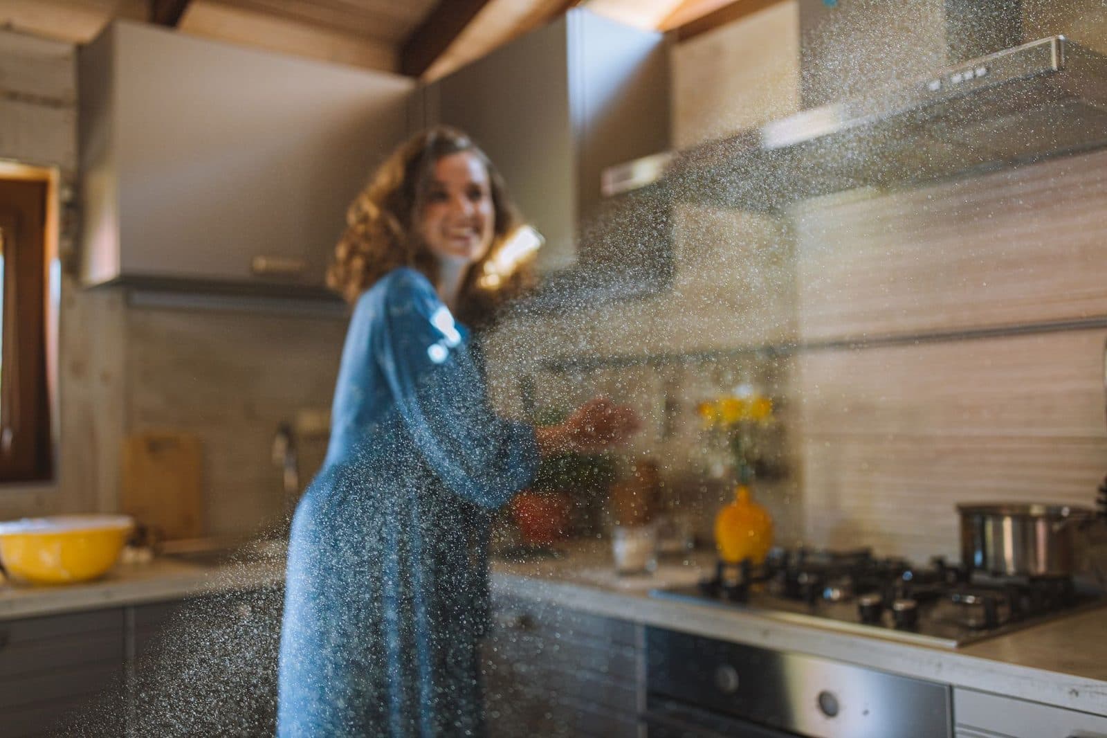 A woman in a blue dress joyfully bakes in a sunlit kitchen, surrounded by cookware and sunbeams.
