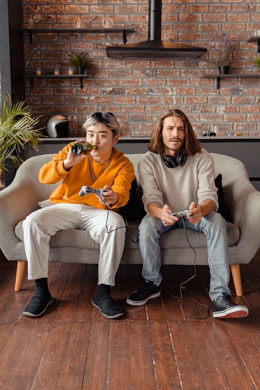 Two friends enjoy gaming and beer on a sofa in an urban apartment setting.