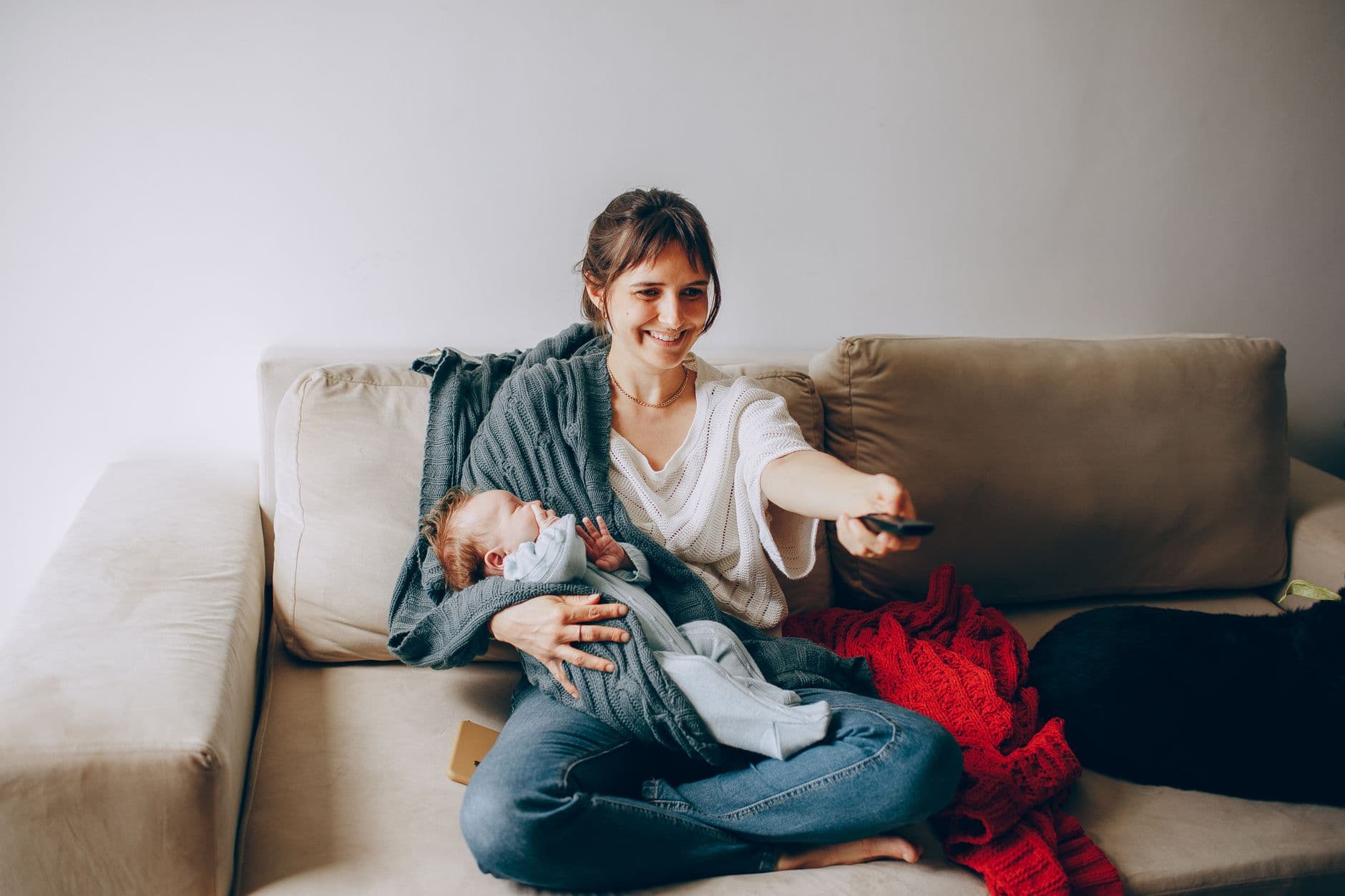 A mother comfortably watches television while holding her sleeping baby on a cozy sofa.