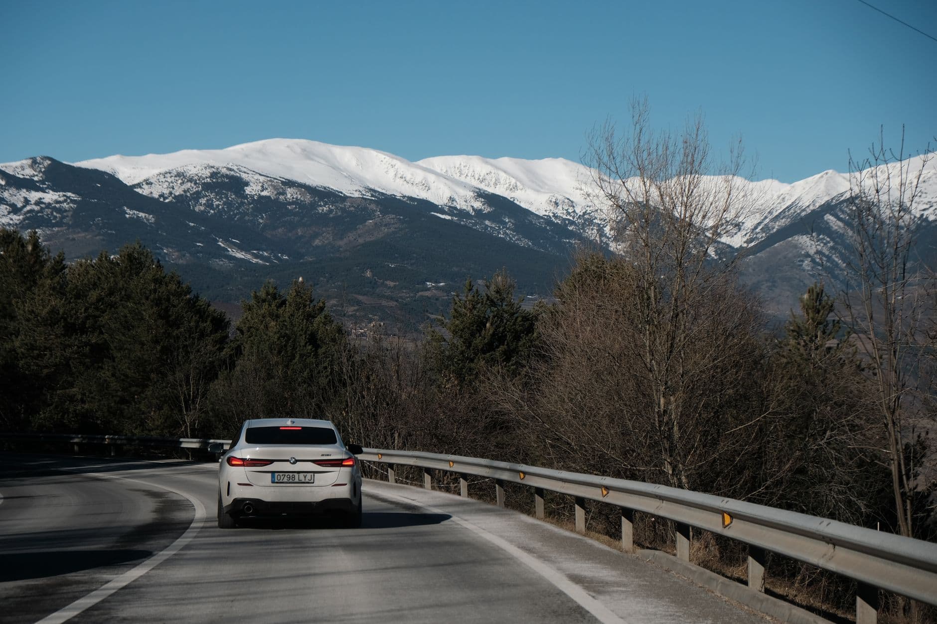 A white car driving on a curvy mountain road with snow-capped peaks in the background.